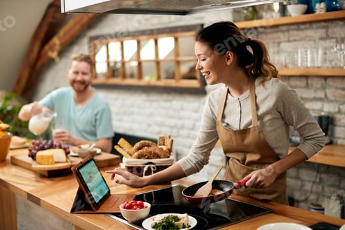Preview: Happy woman using digital tablet while preparing food in the kitchen.