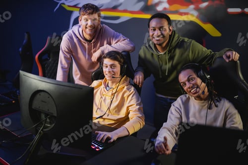 Preview: Positive multicultural friends in headphones looking at camera near computers in gaming club