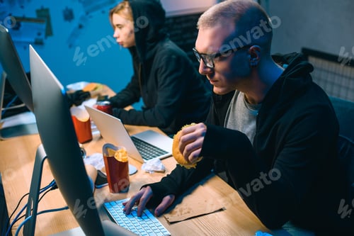 Preview: male hacker working on malware with accomplice and eating junk food