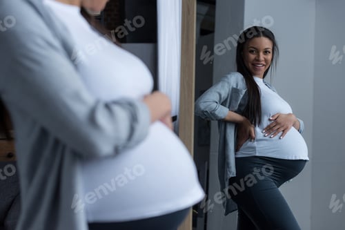 Preview: attractive african american pregnant woman looking at her tummy in mirror