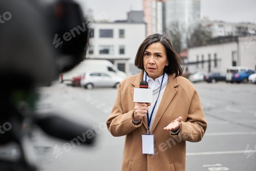Preview: Multiracial journalist in a beige coat reports live amidst an urban backdrop on a cloudy day