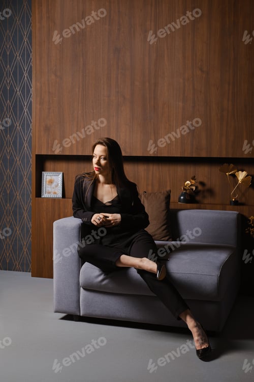 Preview: Portrait of an attractive woman in a black suit sitting in armchair in a modern apartment
