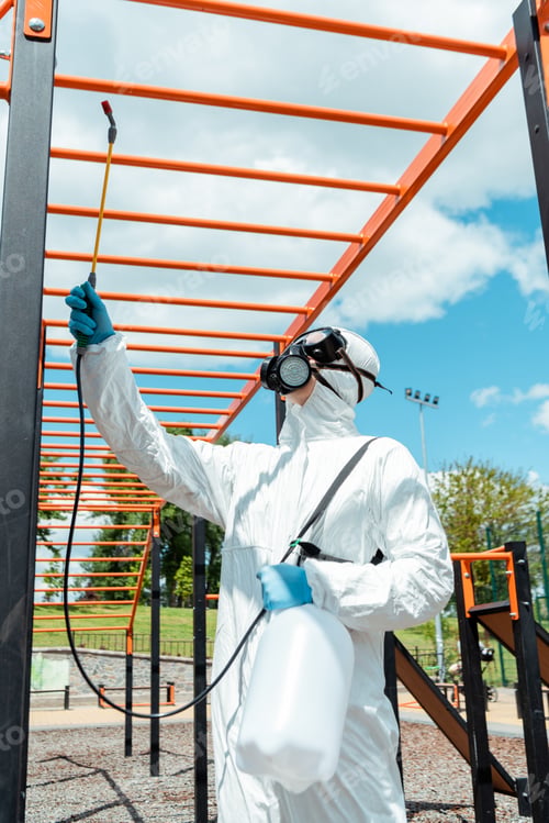Preview: workman in hazmat suit and respirator disinfecting sports ground in park during covid-19 pandemic