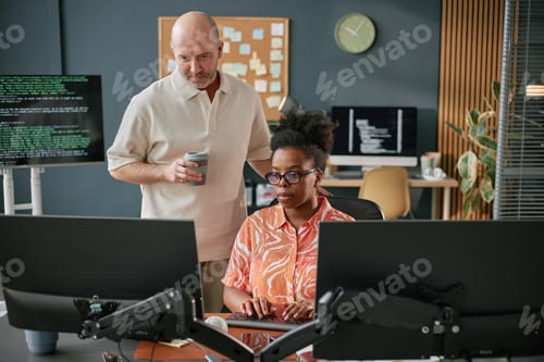 Preview: Young Adult Caucasian Man Supervising Young Adult Black Woman Coding at Workstation