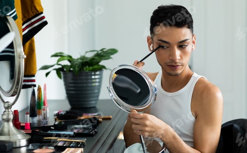Preview: Biracial transgender man looking in mirror and putting on make-up, applying eyeshadow