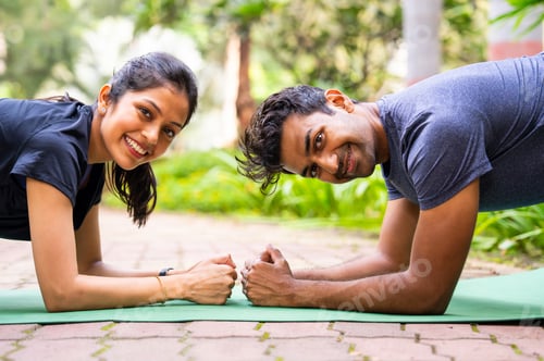 Preview: Indian beautiful young couple doing plank together on mat in outdoor park facing each other
