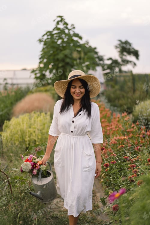 Preview: Beautiful brunette girl in a white dress and hat with watering can watering flowers