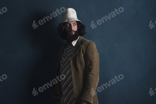 Preview: Retro Fashion Man With Beard Wearing Brown Jacket And White Hat. Leaning Against Gray Wall.