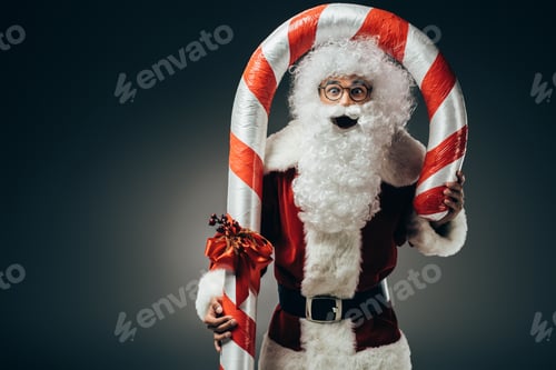 Preview: surprised santa claus in costume standing with big striped christmas stick isolated on grey