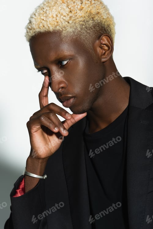 Preview: young african american man with blonde hair posing with hand near face on grey