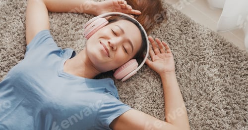 Preview: Young Asian university woman with casual wear headphone lay down on carpet floor.