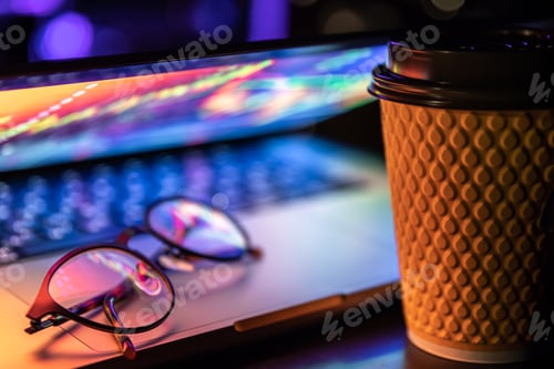 Preview: A laptop half closed in the dark with colourful glow, cup of coffee and glasses.
