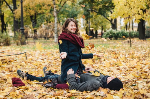 Preview: Positive woman wears knitted scarf, sits on boyfriend, throws yellow leaves in air