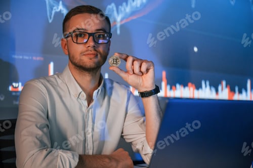 Preview: Holding physical bitcoin in hand. Stock broker trading online in the office with big screen
