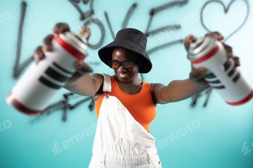 Preview: African Hipster girl painting graffiti on teal wall holding spray bottles wearing orange sunglasses