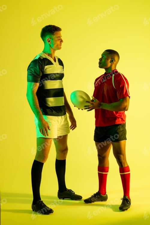 Preview: Diverse male rugby players with rugby ball over yellow lighting