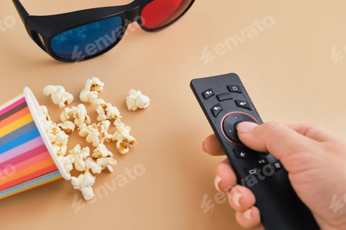 Preview: Hand holds TV remote, bucket of popcorn and 3D glasses. Flat lay, Top view.