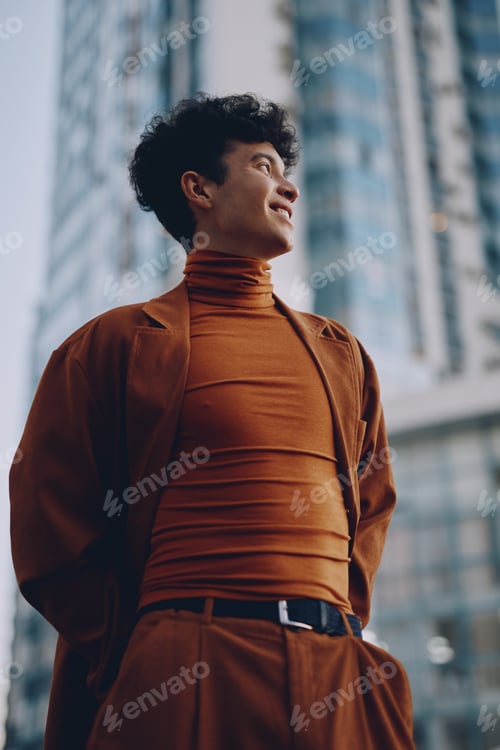 Preview: Portrait of a young man in an elegant orange outfit standing confidently against a modern cityscape