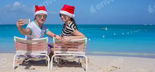 Preview: Young happy couple in red Santa hats taking a photo on cell phone