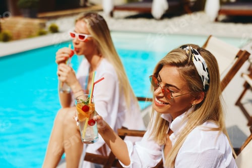 Preview: Two beautiful young women are sitting by the pool and enjoying cocktails