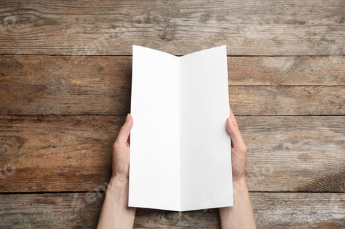 Preview: Young woman holding blank brochure at wooden table, top view. Mock up for design