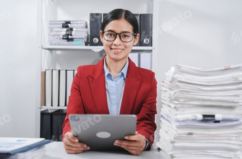 Preview: cheerful individual wearing glasses and a red blazer, hard working with large stack of papers