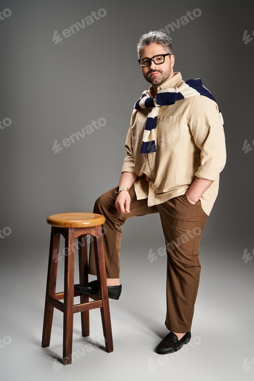 Preview: Stylish man in elegant outfit poses confidently in studio setting with wooden stool