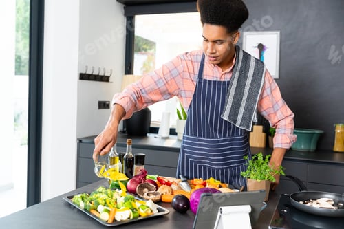 Preview: Happy biracial man wearing apron cooking dinner, pouring vegetables in kitchen, unaltered