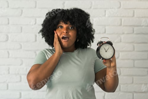 Preview: Surprised african american woman holding alarm clock amazed with open mouth and surprise happy face