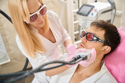 Preview: Blonde woman performs laser hair removal procedure on young mans face