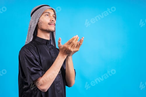 Preview: Side View Of Young Male Wearing Arabian Style Raising Hand To Pray Isolated On Blue Background