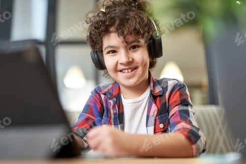 Preview: Cheerful latin american school boy wearing headphones, smiling at camera and making notes. Child