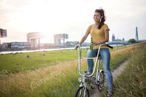 Preview: Germany, Cologne, young woman with bicycle on meadow near Rhine River at evening twilight