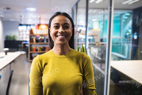 Preview: Woman smiling at camera in modern open-plan office, with desks, monitors and potted plants