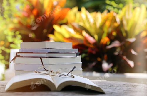 Preview: Open book with eyeglasses and pile books on table closeup outdoor nature blur background