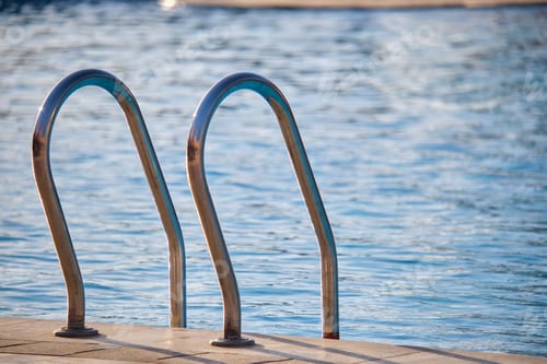 Preview: Close up of swimming pool stainless steel handrail descending into tortoise clear pool water.