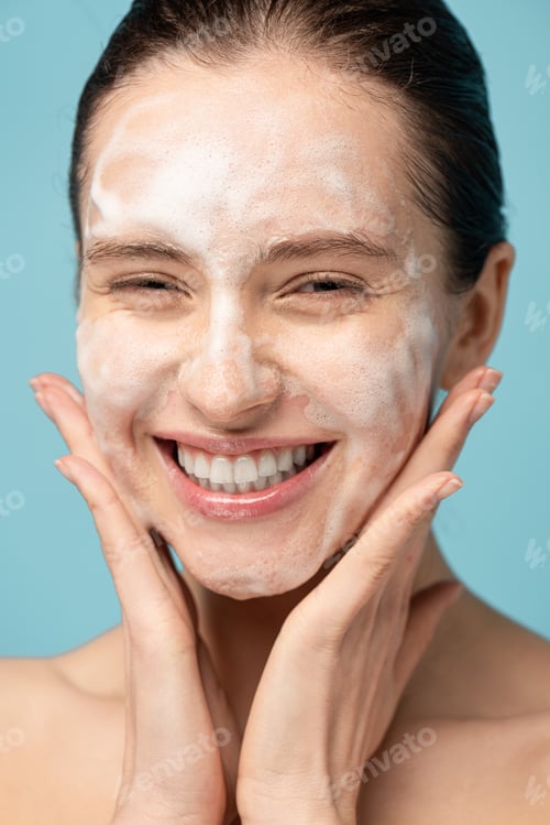 Preview: beautiful smiling young woman applying cleansing foam on face, isolated on blue