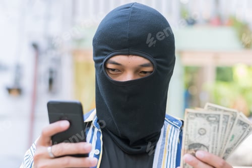 Preview: man close up thief in a mask and colorful shirt on blurred background holding the US dollars money