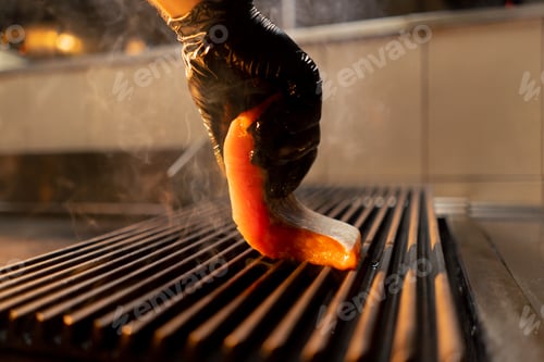 Preview: close-up of a chef's hand in a glove puts a raw piece of salmon on the grill in a restaurant kitchen