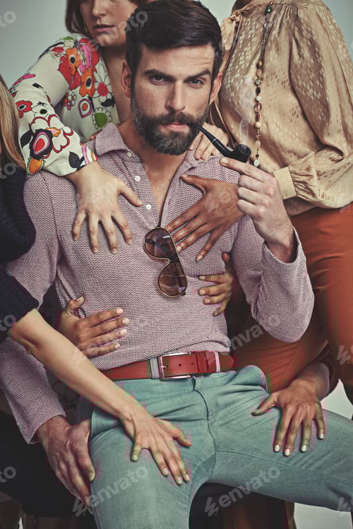 Visualização: Foto de estúdio de um homem atraente usando roupas retrô dos anos 70 sendo tocado por mulheres enquanto fuma cachimbo