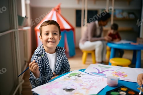 Preview: Happy kid drawing with watercolor paints at preschool and looking at camera.