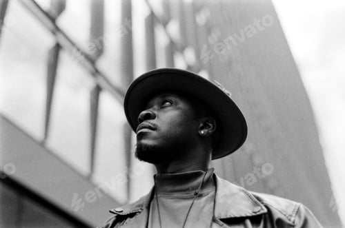 Vista previa: Dramatic black and white portrait of man wearing a hat looking up in front of building.