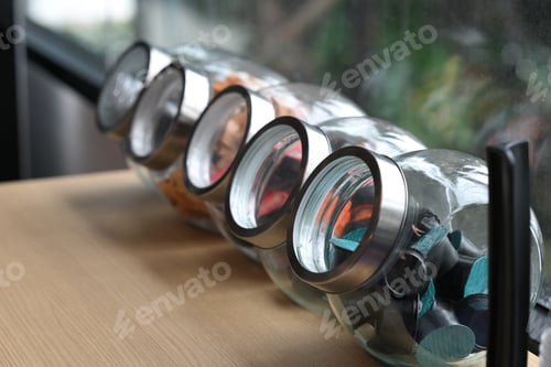 Preview: Close up view of glass jars filled with colorful coffee capsules on a wooden counter by the window