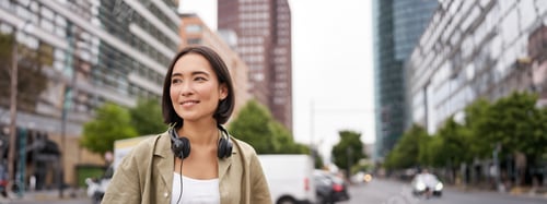 Preview: Portrait of young asian woman in headphones, posing in city, smiling and looking away, standing on
