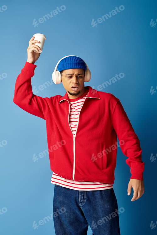 Preview: Trendy young african american man enjoying music with coffee against blue backdrop