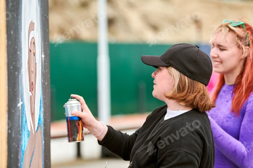 Visualização: Duas artistas femininas pintando quadros com lata de tinta spray pulverizando sobre tela em exposição de rua
