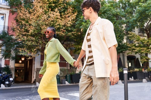 Preview: Stylish diverse couple holding hands and walking on street