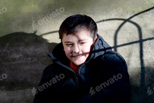 Preview: Happy boy child is smiling sitting next to old wall enjoying relaxing in sunny day spring park