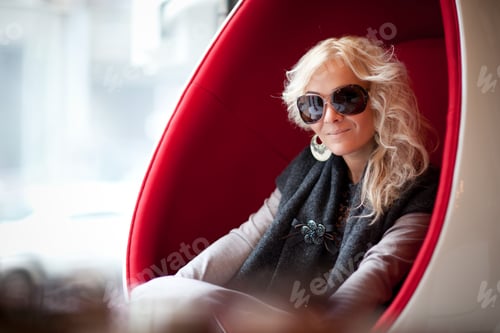 Preview: Young beautiful blond woman in big sunglasses sitting in red egg shaped armchair