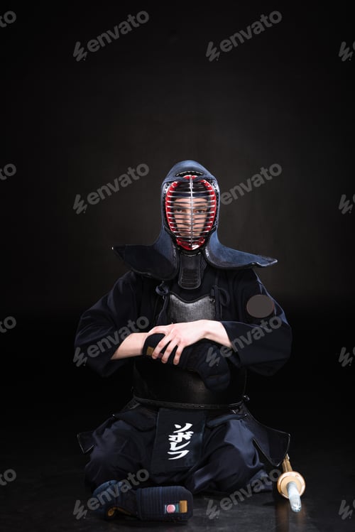 Preview: Kendo fighter in helmet sitting on floor and taking off glove on black